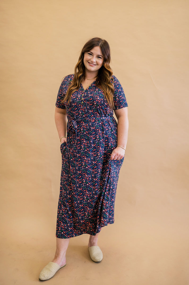 Model smiling while styled in a comfortable fit, navy floral print dress. The dress is a modest design with full coverage for all bodies, has a mid calf length, short sleeves, and a removable tie. Styled with tan slides and gold jewelry the ditsy floral dress can be worn both casually and dressed up easily. 
