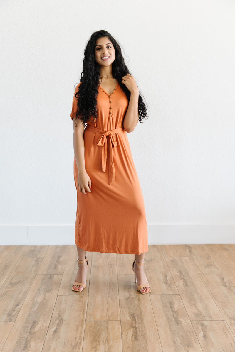 Model wearing the MW Anywhere dress in Rust. Dress has buttons, a waist tie, and comes to mid thigh.