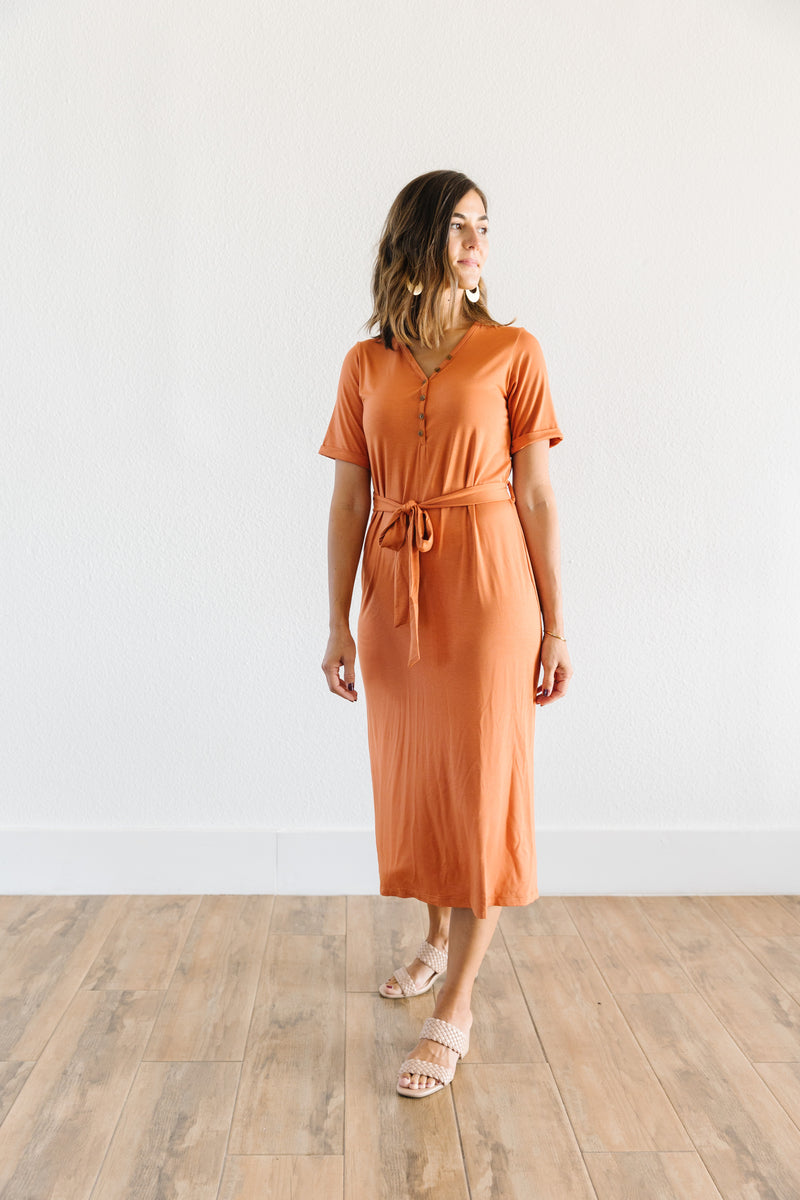 Model in the MW Anywhere dress in Rust. Dress has buttons, a waist tie, and comes to mid thigh.