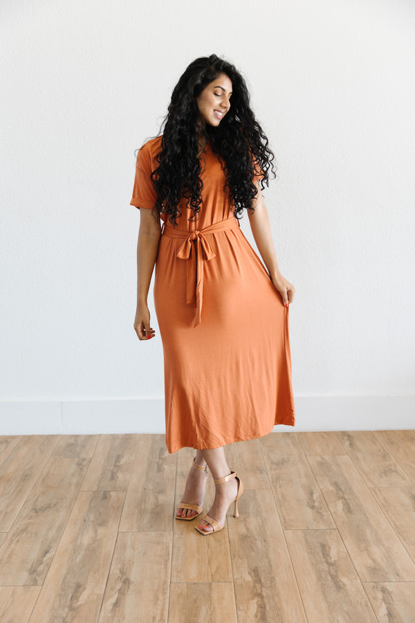 Model wearing the MW Anywhere dress in Rust. Dress has buttons, a waist tie, and comes to mid thigh.