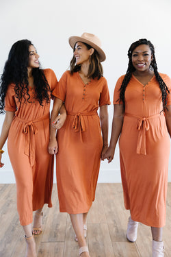 Three women wearing the MW Anywhere dress in Rust. Dress has buttons, a waist tie, and comes to mid thigh.