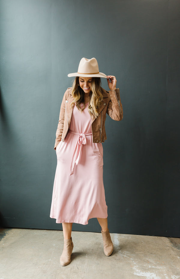 Model wearing dusty pink anywhere dress with booties, jacket and hat. Dres has a v-neck design, is a light dusty pink color, is the perfect midi length and has a removable waist tie.